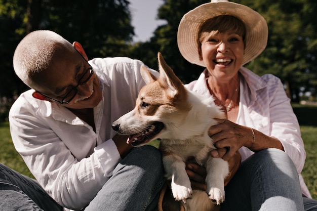 Uomo dai capelli grigi in occhiali e camicia bianca sorridente e in posa con corgi carino e donna bionda con cappello e camicetta leggera all'aperto