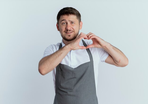 Uomo del barbiere in grembiule che fa il gesto del cuore con le dita sorridendo allegramente in piedi su sfondo bianco