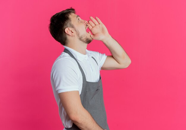 Uomo del barbiere in grembiule che grida o chiama qualcuno con la mano vicino alla bocca in piedi su sfondo rosa