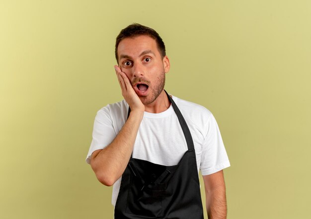 Uomo del barbiere in grembiule che guarda in avanti con la mano sulla sua guancia stupito e sorpreso in piedi sopra la parete chiara