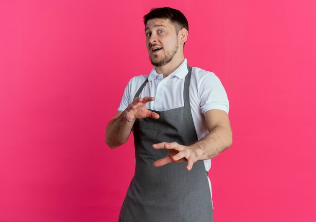 Uomo del barbiere in grembiule che guarda l'obbiettivo con espressione disgustata che fa il gesto di difesa con le mani in piedi su sfondo rosa