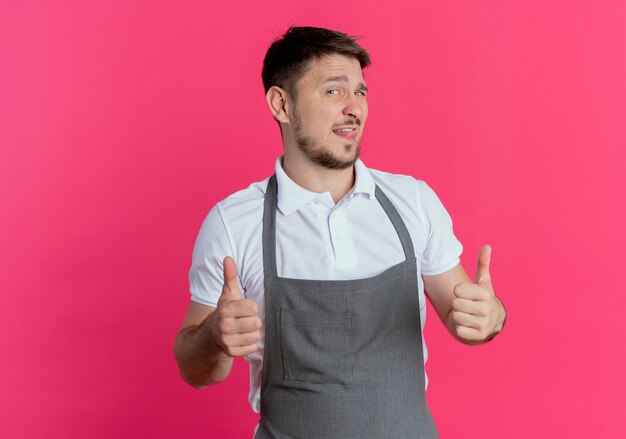 Uomo del barbiere in grembiule che guarda l'obbiettivo sorridente che mostra i pollici in su in piedi su sfondo rosa