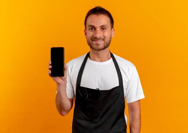 Uomo del barbiere in grembiule che mostra smartphone sorridente amichevole in piedi sopra la parete arancione