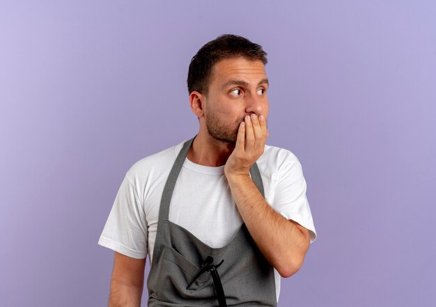 Uomo del barbiere in grembiule che osserva da parte ha sottolineato e nervoso che copre la bocca con la mano in piedi sul muro viola