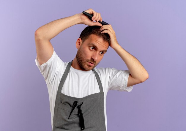 Uomo del barbiere in grembiule che pettina i capelli con la faccia seria in piedi sopra il muro viola