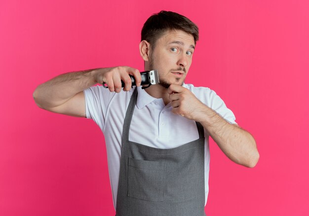 Uomo del barbiere in grembiule che taglia la sua barba che guarda l'obbiettivo in piedi su sfondo rosa