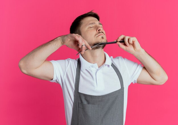 Uomo del barbiere in grembiule che taglia la sua barba con le forbici in piedi su sfondo rosa