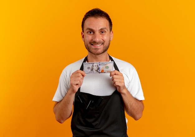 Uomo del barbiere in grembiule che tiene contanti guardando in avanti con il sorriso sul viso in piedi sopra la parete arancione