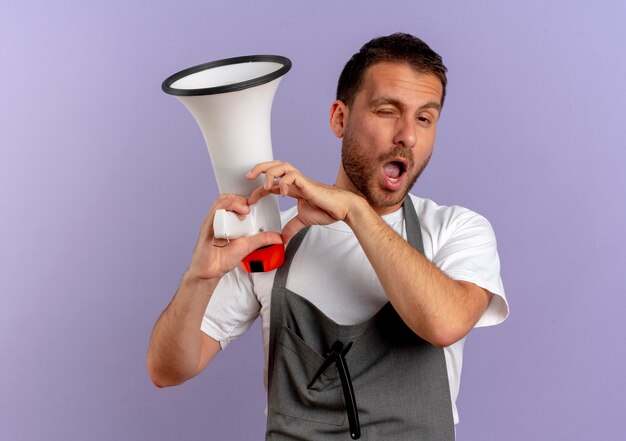 Uomo del barbiere in grembiule che tiene il megafono guardando in avanti sorridendo e ammiccando facendo il gesto del cuore con le dita sul petto in piedi sopra il muro viola