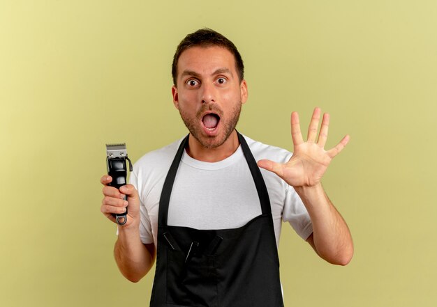 Uomo del barbiere in grembiule che tiene la macchina per il taglio dei capelli guardando in avanti sorpreso con la mano alzata aperta in piedi sopra il muro leggero