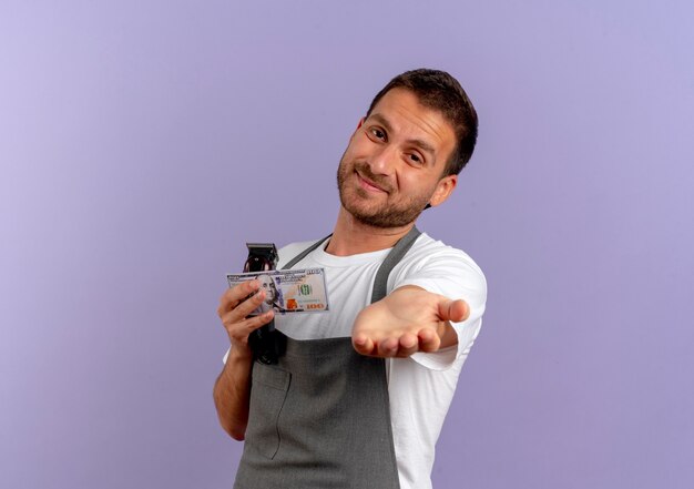 Uomo del barbiere in grembiule che tiene la macchina per il taglio dei capelli tenendo la mano davanti a sé per chiedere soldi in piedi sopra il muro viola