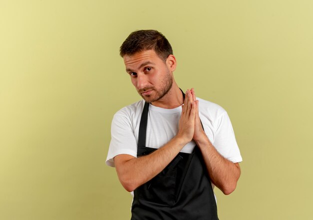 Uomo del barbiere in grembiule che tiene le palme insieme guardando in avanti con la faccia seria in attesa di qualcosa in piedi sopra la parete leggera