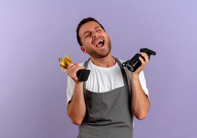 Uomo del barbiere in grembiule che tiene macchina per il taglio dei capelli e trofeo felice ed eccitato in piedi sopra la parete viola