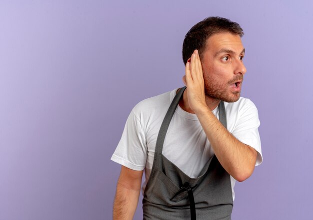 Uomo del barbiere in grembiule che tiene macchina per il taglio dei capelli tenendo la mano vicino al suo orecchio cercando di ascoltare in piedi sopra il muro viola