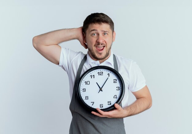 Uomo del barbiere in grembiule che tiene orologio da parete che guarda l'obbiettivo perplesso e confuso in piedi su sfondo bianco