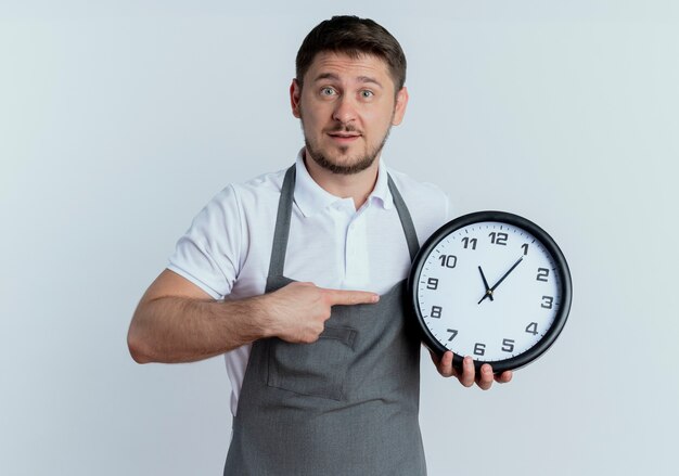 Uomo del barbiere in grembiule che tiene orologio da parete che indica con il dito che sembra sorpreso in piedi su sfondo bianco