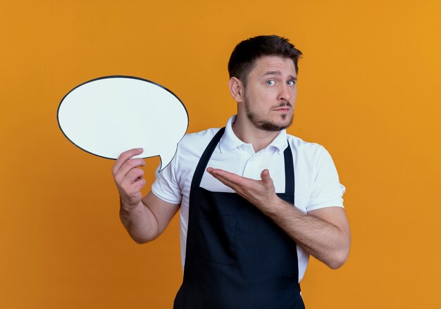 Uomo del barbiere in grembiule che tiene segno in bianco della bolla di discorso che presenta con il braccio della sua mano che sta sopra fondo arancio