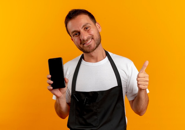 Uomo del barbiere in grembiule che tiene smartphone sorridendo allegramente mostrando i pollici in su guardando in avanti in piedi sopra la parete arancione