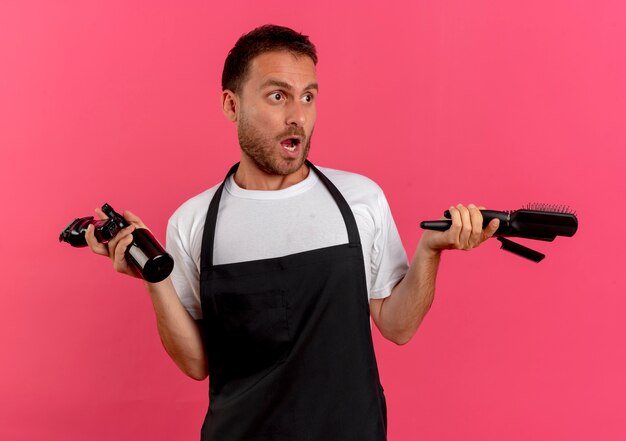 Uomo del barbiere in grembiule che tiene spray con spazzola per capelli d'acqua e trimmer guardando da parte confuso in piedi sopra il muro rosa