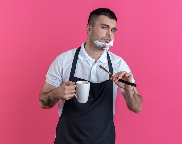 Uomo del barbiere in grembiule con schiuma da barba sul viso andando a radersi con il rasoio guardando la telecamera sorridente fiducioso in possesso di una tazza in piedi su sfondo rosa