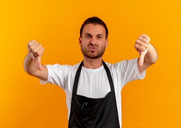 Uomo del barbiere in grembiule guardando in avanti dispiaciuto che mostra i pollici verso il basso in piedi sopra la parete arancione