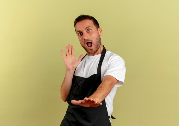 Uomo del barbiere in grembiule guardando in avanti tenendo le braccia fuori in piedi sorpreso e felice sopra la parete chiara