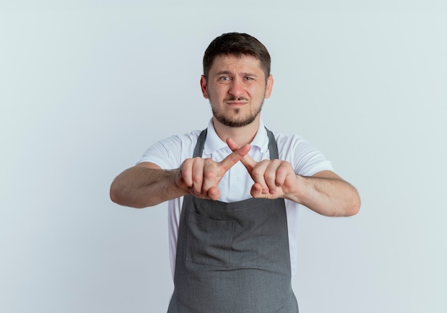 Uomo del barbiere in grembiule scontento che mostra il segnale di stop che attraversa le dita indice in piedi sul muro bianco