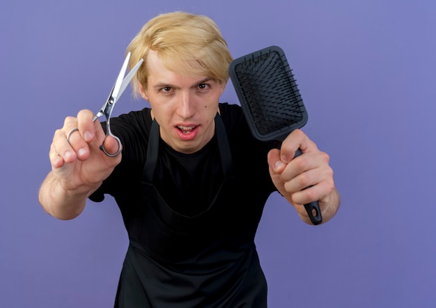 Uomo del barbiere professionista in grembiule che mostra le forbici e la spazzola per capelli guardando la parte anteriore con la faccia seria in piedi sopra la parete blu