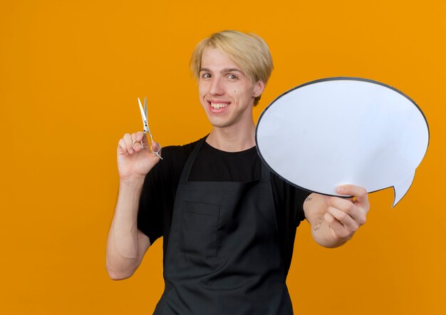 Uomo del barbiere professionista in grembiule che tiene il segno in bianco della bolla di discorso e le forbici che esaminano la parte anteriore che sorride con la faccia felice che sta sopra la parete arancione