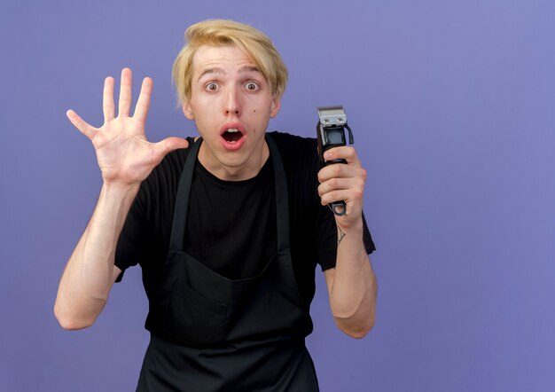 Uomo del barbiere professionista in grembiule che tiene il trimmer guardando davanti sorpreso e stupito mostrando il numero cinque in piedi sopra la parete blu