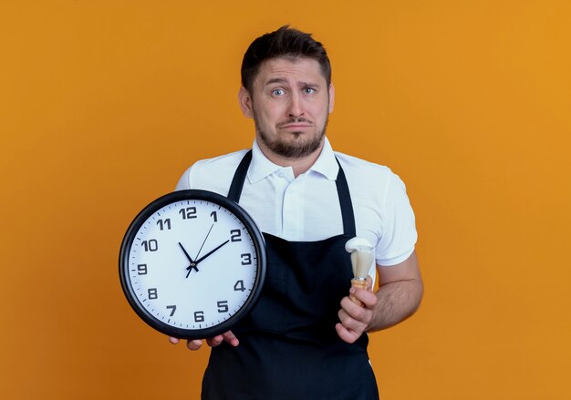 Uomo del barbiere sconvolto in grembiule che tiene orologio da parete e pennello da barba che guarda l'obbiettivo con espressione triste in piedi su sfondo arancione