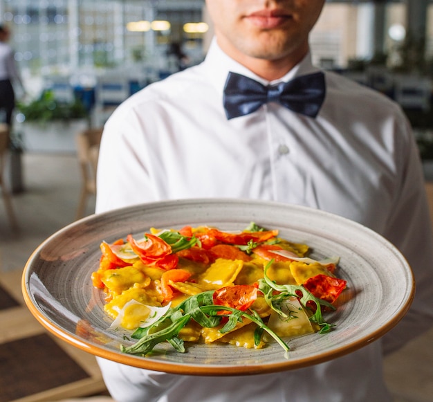 Uomo del cameriere che tiene un grande piatto di ravioli con foglie di rucola, parmigiano