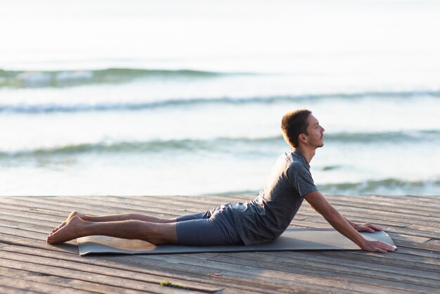 Uomo del colpo completo che pratica posa di yoga vicino al mare