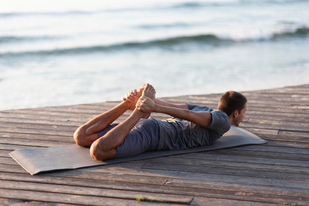 Uomo del colpo completo che pratica posa di yoga vicino al mare