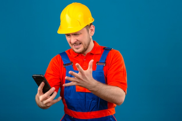 Uomo del costruttore che indossa l'uniforme da costruzione e l'elmetto di sicurezza essendo arrabbiato confuso aggressivo di cattivo umore dopo aver sentito cattive notizie sulla telefonata che urla minaccioso nell'altoparlante del suo smartphone