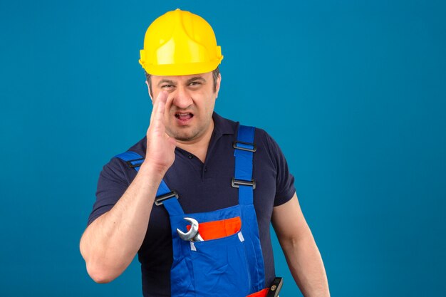 Uomo del costruttore che indossa l'uniforme della costruzione e casco di sicurezza che urla qualcosa e che tiene la mano vicino alla sua bocca aperta sopra la parete blu isolata