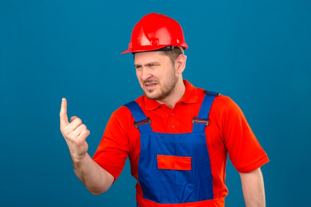 Uomo del costruttore che indossa l'uniforme edile e casco di sicurezza che indica scontento e frustrato al lato che controlla la parete blu isolata