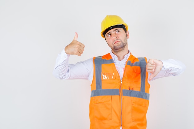 Uomo del costruttore che mostra i pollici su e giù in camicia, uniforme e pensieroso, vista frontale.