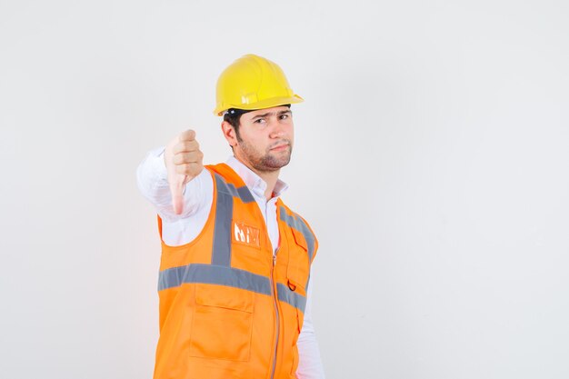 Uomo del costruttore che mostra il pollice giù in camicia, uniforme e che sembra scontento. vista frontale.