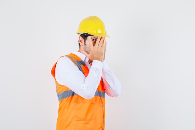 Uomo del costruttore che osserva attraverso le dita con un occhio in camicia, uniforme e che sembra imbarazzato.