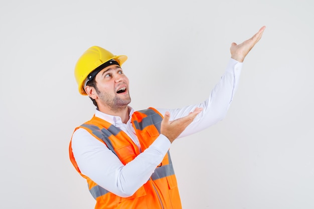 Uomo del costruttore che osserva in su mentre mostra qualcosa in camicia, uniforme e che sembra allegro. vista frontale.