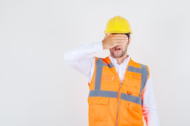 Uomo del costruttore che tiene la mano sugli occhi in camicia, uniforme, vista frontale.