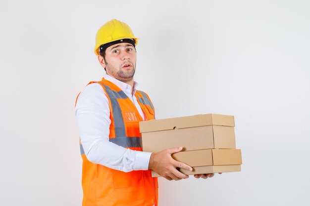 Uomo del costruttore che tiene scatole di cartone in camicia, uniforme e dall'aspetto irresoluto. vista frontale.