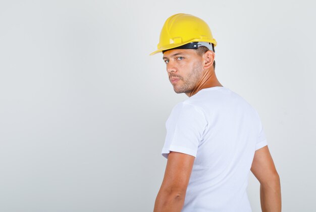 Uomo del costruttore girando e guardando indietro in t-shirt bianca, casco e guardando serio, vista posteriore.
