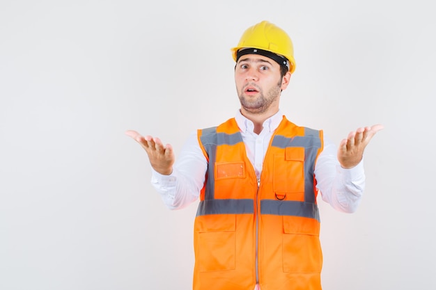 Uomo del costruttore in camicia, uniforme che alza le braccia nel gesto interrogativo e che sembra perplesso, vista frontale.