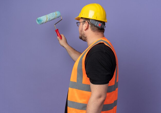 Uomo del costruttore in giubbotto da costruzione e casco di sicurezza che dipinge un muro con rullo di vernice in piedi su sfondo blu