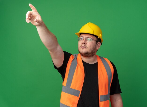 Uomo del costruttore in giubbotto da costruzione e casco di sicurezza che guarda in alto con una faccia seria che punta con il dito indice verso qualcosa che sta su sfondo verde