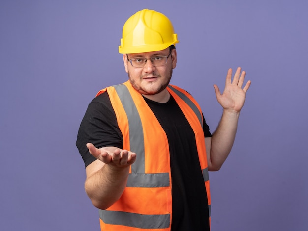 Uomo del costruttore in giubbotto da costruzione e casco di sicurezza che guarda l'obbiettivo con le braccia fuori sorridente fiducioso in piedi su sfondo blu