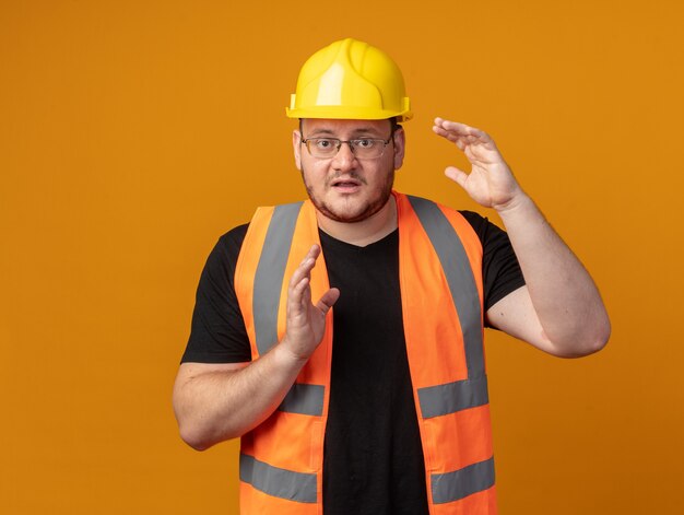 Uomo del costruttore in giubbotto da costruzione e casco di sicurezza che guarda la telecamera confusa che mostra il gesto delle dimensioni con le mani preoccupate in piedi su sfondo arancione