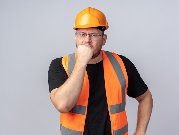 Uomo del costruttore in giubbotto da costruzione e casco di sicurezza che guarda la telecamera stressato e nervoso che si morde le unghie in piedi sopra il bianco